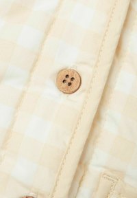 Tessuto beige chiaro con motivo a scacchi, caratterizzato da un bottone in legno con quattro fori, dettaglio cucito lungo i bordi e superficie liscia.