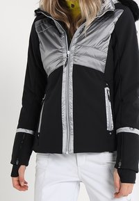 Schwarze und graue Skijacke mit wattiertem Design, versehen mit einem pelzgefütterten Kapuzen, silbernen Reißverschlüssen und Reißverschlusstaschen zur Aufbewahrung.