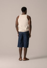 Débardeur en maille beige clair et shorts en denim bleu foncé avec deux poches arrière, accompagnés de sandales marron. Fond uni.
