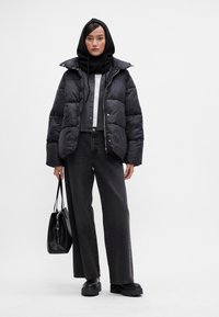 Casaco puffer preto com colarinho alto, combinado com uma camisa cinza e jeans pretos de perna larga. Segura uma bolsa preta e usa um hoodie preto.