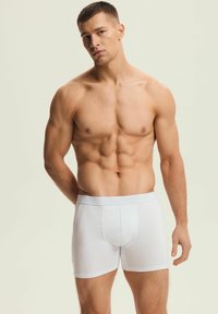 Białe bawełniane bokserki z miękką elastyczną gumką w talii, na której widnieje nazwa marki. Model stoi z neutralnym wyrazem twarzy na jasnym tle.