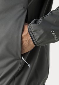 Grijze jas met een gladde textuur, voorzien van een ritszak en een elastische manchet met "TEXASHIELD" erop gedrukt. Koorddetail aan de zoom.