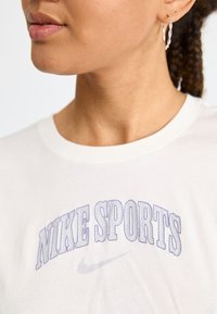 T-shirt en coton blanc avec le texte brodé "NIKE SPORTS" en violet et un petit logo Nike swoosh gris en dessous. Texture lisse.
