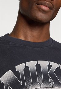 Tmavě modré bavlněné tričko s velkým texturovaným bílým potiskem "NIKE" na hrudi, s žebrovaným kulatým výstřihem a hladkým materiálem.