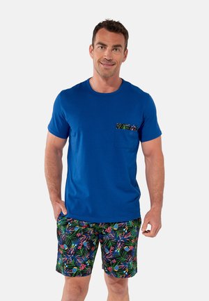 Mann trägt ein blaues T-Shirt mit einer gemusterten Tasche und passenden Shorts mit tropischem Blumenmuster, steht mit einer Hand in der Hosentasche.
