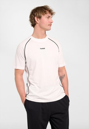 Blond man with short hair and mustache wearing a white short-sleeve shirt with black piping and black pants, standing with hands in pockets.