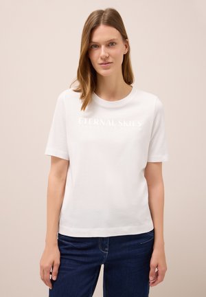Donna con capelli castano chiaro che indossa una maglietta bianca a maniche corte con la scritta "ETERNAL SKIES" e jeans blu scuro, su uno sfondo beige semplice.