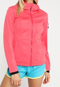 Pink zip-up jacket with a fitted design, soft texture, and hood; features black accents and zippered side pockets. Worn with blue shorts.
