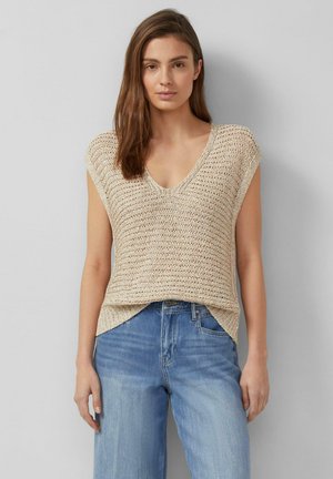 Kobieta ubrana w beżowy, dziergany, bezrękawnik z dekoltem w serek, zestawiony z jasnoniebieskimi, wysokimi dżinsami, stoi na szarym tle.