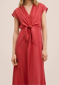 Femme portant une robe rouge sans manches avec un décolleté en V, des boutons sur le devant et un nœud noué à la taille.