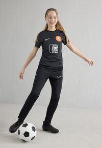 Meisje in een zwart voetbaltenue die een zwart-witte voetbal trapt op een egale lichtgrijze vloer met een bijpassende achtergrond.