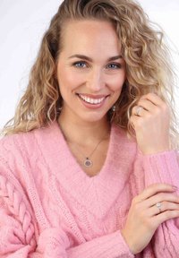 Roze gebreide V-hals trui met kabelpatronen, zilveren ketting met een ronde hanger en een zilveren ring aan de rechterhand.