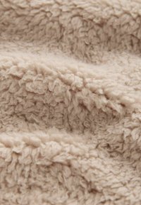 Beige fleece-stof med en blød, tekstureret overflade, der har tætte, krøllede fibre, hvilket skaber et varmt og hyggeligt udseende.
