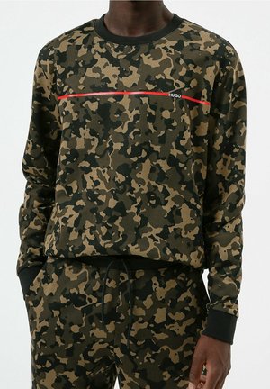 Mężczyzna w zielono-czarnej kamuflażowej bluzie z długim rękawem i dopasowanych spodniach, z cienką czerwoną linią i napisem "HUGO" na piersi.