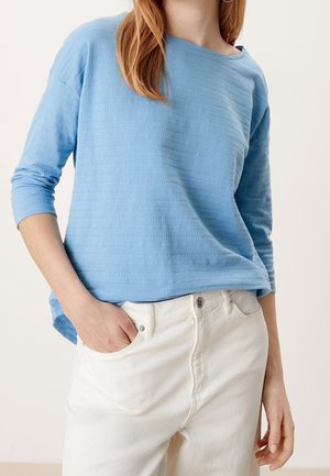 Haut à manches longues bleu clair texturé, porté avec un pantalon taille haute blanc cassé, une main dans la poche, visible du cou aux hanches.