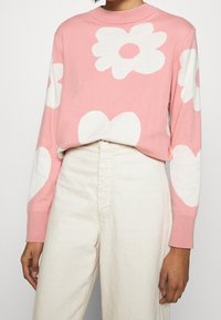 Ljusrosa tröja med vita blommor och hjärtmönster, ribbad rund halsringning och långa ärmar, kombinerad med off-white höga byxor.