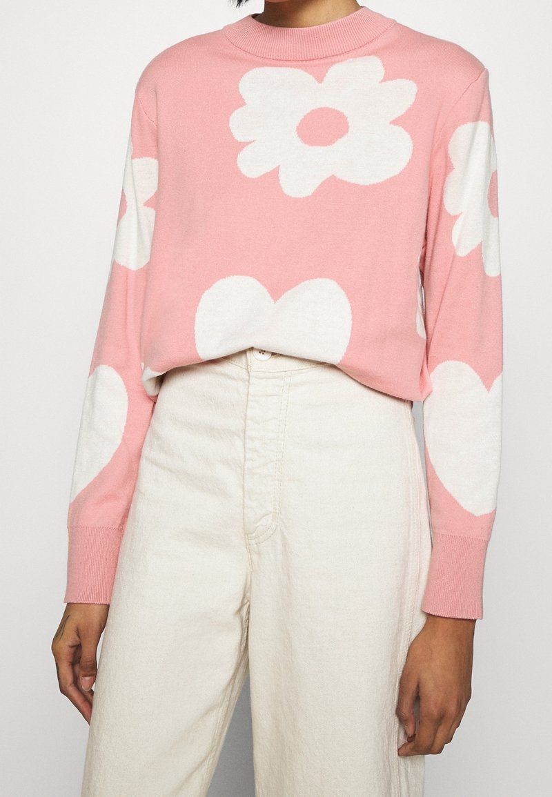Ljusrosa tröja med vita blommor och hjärtmönster, ribbad rund halsringning och långa ärmar, kombinerad med off-white höga byxor.