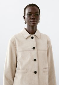 Beige mockaskinnjacka med krage, framknäppning och två sidofickor. Har svarta knappar och diskret sömnadsdetalj.