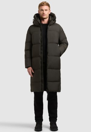Man die een lange, donkere donsjas met capuchon, zwarte broek en zwarte laarzen draagt, staand tegen een simpele lichte achtergrond.