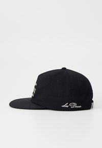 Black cap with a curved brim, made of fabric, features white embroidered text and branding on the front and side. Casual design.