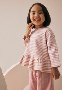 Tricou roz deschis cu mânecă lungă și tiv peplum, cu un model pătrățos, asortat cu colanți roz cu textură ribbată. Textură netedă, design casual.