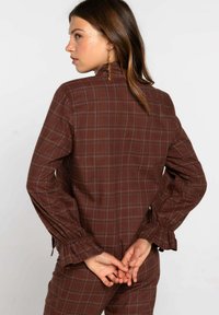 Camisa de cuadros marrón de manga larga, con puños acampanados y cuello alto. Presenta una tela texturizada y un patrón de cuadros clásico.