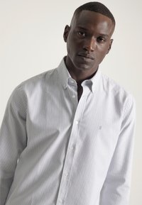Camisa de botones a rayas en gris claro y blanco, con un cuello clásico, mangas largas y un pequeño logo en el pecho. Textura de tela suave.