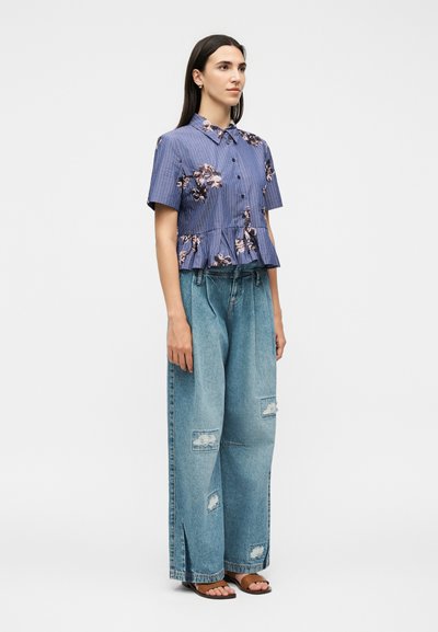 Femme aux longs cheveux foncés portant un haut bleu à fleurs à manches courtes boutonné et un jean en denim délavé à jambes larges avec des sandales, debout de côté.