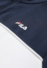 Vêtement zippé bleu marine et blanc avec logo FILA brodé en blanc et rouge sur un panneau en tissu bleu marine.