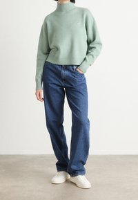 Pull à col roulé en tricot vert menthe avec un ourlet côtelé, associé à un jean bleu ample et des baskets blanches, se tenant sur un fond neutre.