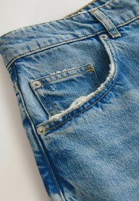 Primer plano del bolsillo de unos vaqueros de mezclilla azul con detalles desgastados, costuras visibles y remaches metálicos sobre un fondo claro.