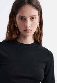 Schwarzer Strickpullover mit Rundhalsausschnitt und gerippten Details an den Schultern. Das Modell trägt silberne Creolen; der Hintergrund ist einfarbig weiß.