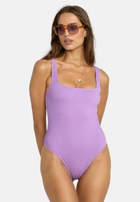 Maillot de bain une pièce violet à côtes avec des bretelles épaisses et texturées ainsi qu'un décolleté carré, associé à des lunettes de soleil aux tons ambre et un collier en or.