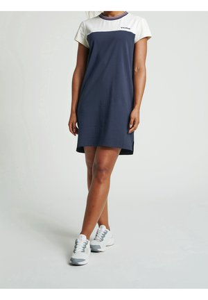 Frau trägt ein kurzärmeliges, marineblau-weißes Freizeitkleid mit Sneakers und steht vor einem einfarbigen hellen Hintergrund.