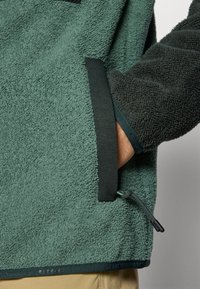 Flis jakna u tamnozelenoj boji s teksturiranim materijalom, kontrastnim crnim detaljima i podesivim kabelom na rubu za sigurno prilaganje.