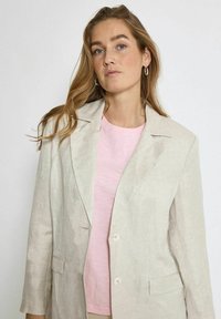 Šviesiai béžinis lininis švarkas su vienu mygtuku, lengvai tekstūruota medžiaga ir dviem priekiniams kišenėms, derinamas su minkštos rožinės spalvos marškiniais po juo.