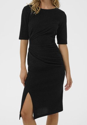 Kobieta w dopasowanej czarnej sukience midi z rękawami do łokcia, bocznym rozporkiem oraz marszczeniami na wysokości talii i bioder.