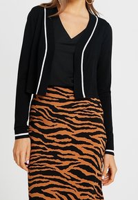 Schwarze, verkürzte Strickjacke mit weißen Akzenten, kombiniert mit einem schwarzen Oberteil und einem rock mit braunem Streifen und Tigerprint. Der Stoff ist weich und hat eine glatte Textur.