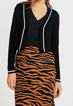 Cardigan noir court avec des accents blancs, associé à un haut noir et une jupe à imprimé tigre rayé marron. Le tissu est doux avec une texture lisse.