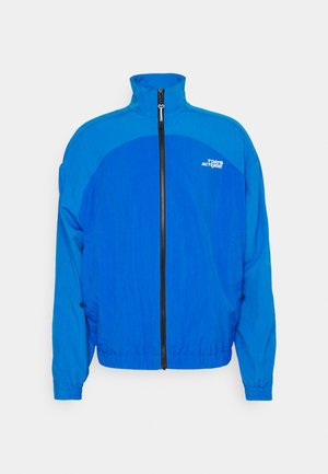 Kék széldzseki, magas gallérral, teljes cipzárral, rugalmas mandzsettákkal és húzózsinóros aljjal. Kontrasztos paneldesign és embléma a mellkason.