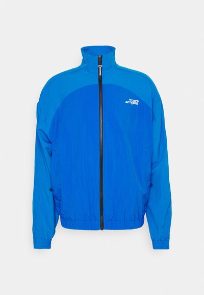 Kék széldzseki, magas gallérral, teljes cipzárral, rugalmas mandzsettákkal és húzózsinóros aljjal. Kontrasztos paneldesign és embléma a mellkason.