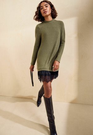 Mujer caminando en interiores con un suéter largo verde oliva, falda negra de encaje, botas negras hasta la rodilla y sosteniendo un pequeño bolso negro.