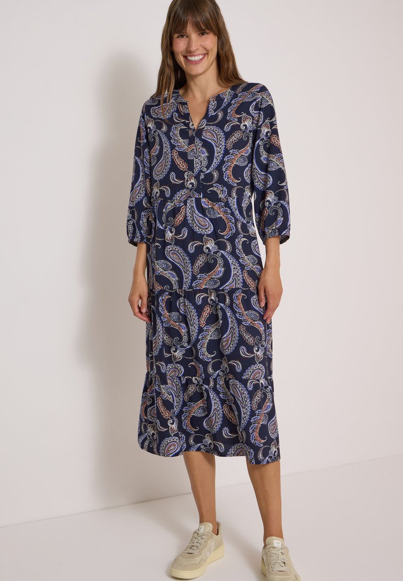 Femme souriante, portant une robe midi bleu marine à motif cachemire avec des manches trois-quarts et des baskets beige, debout contre un mur clair uni.