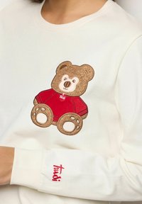 Biała bluza z haftowanym na przodzie brązowym misiem w czerwonej koszulce. Nazwa marki wyhaftowana na mankiecie na czerwono.