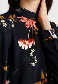 Blusa floreale in tessuto navy scuro con vivaci fiori rossi, arancioni e bianchi. Collo alla coreana e maniche lunghe con dettagli floreali.