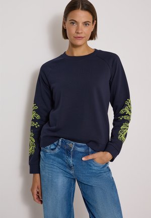 Femme portant un sweat-shirt bleu marine à manches longues avec des motifs floraux verts vifs sur les manches et un jean bleu, debout devant un fond uni.