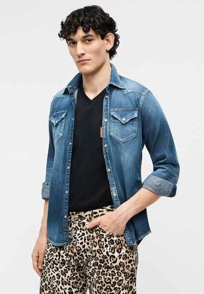 Νέος άνδρας με τζιν πουκάμισο με μανίκια γυρισμένα πάνω από μαύρο μπλουζάκι και παντελόνι με λεοπάρ σχέδιο, στέκεται μπροστά σε απλό ανοιχτό φόντο.