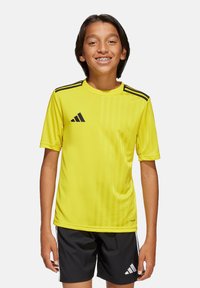 Geel Adidas voetbalshirt met korte mouwen, zwarte drie-streepdashouderaccenten en een verticale streepstructuur. G gedragen met zwarte shorts.
