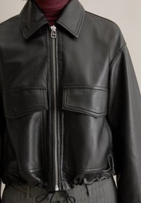 Schwarze Leder-geschnittene Jacke mit Kragen, silbernem Reißverschluss, zwei Fronttaschen und gerafftem Saum. Glatte Textur mit subtiler Schimmer.