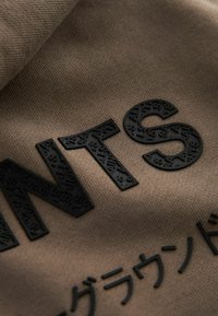 Smeđi materijal s teksturiranim crnim slovima "PANTS" i dodatnim crnim tekstom, koji sadrži složene uzorke na slovima.